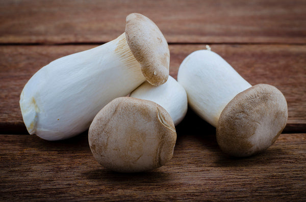 A close-up of fresh king trumpet mushrooms with thick white stems and small tan caps resting on a wooden surface.